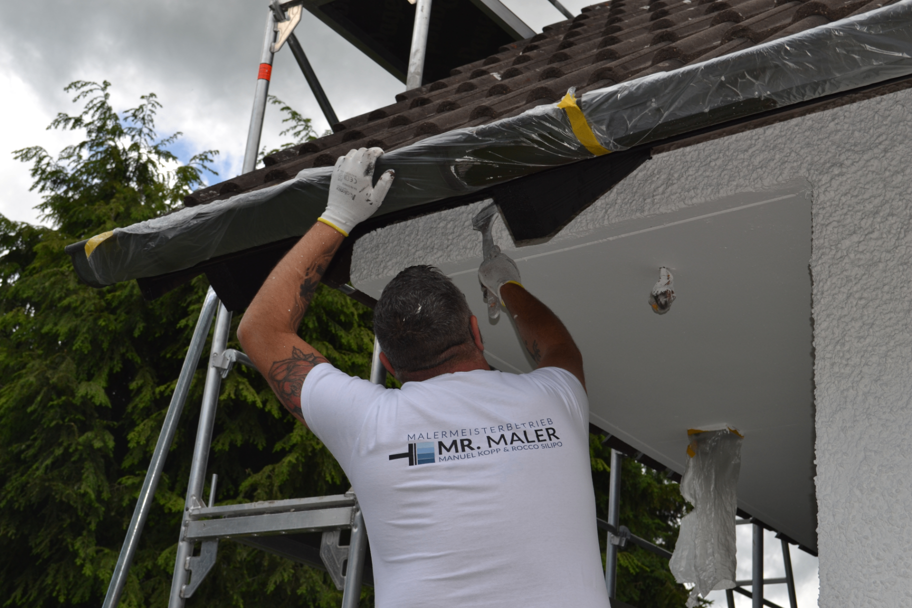 Maler von Mr. Maler beim Abkleben und Streichen eines Dachüberstands auf dem Gerüst